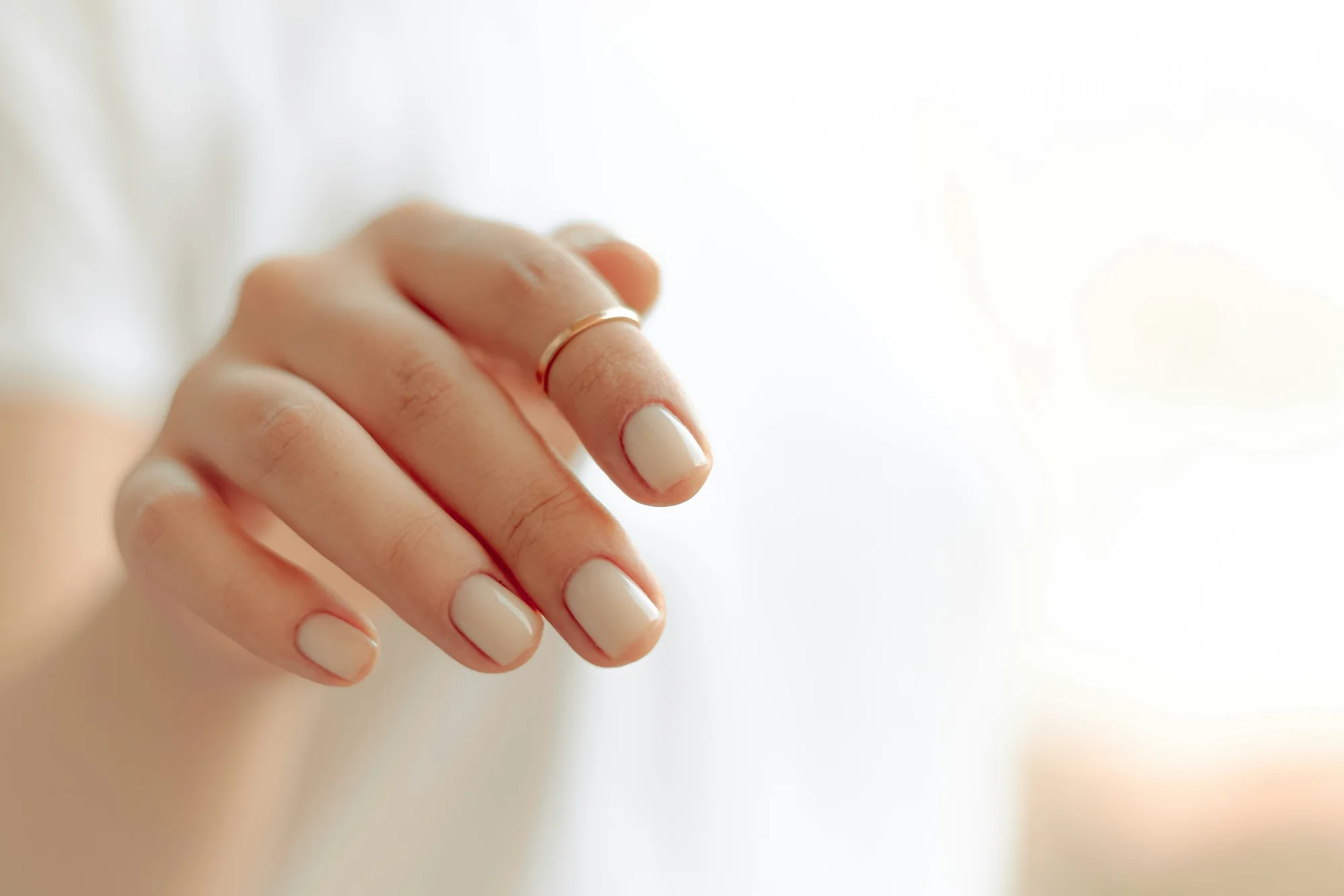 Close-up of a hand with a glossy nude-beige manicure and a thin gold ring against soft white