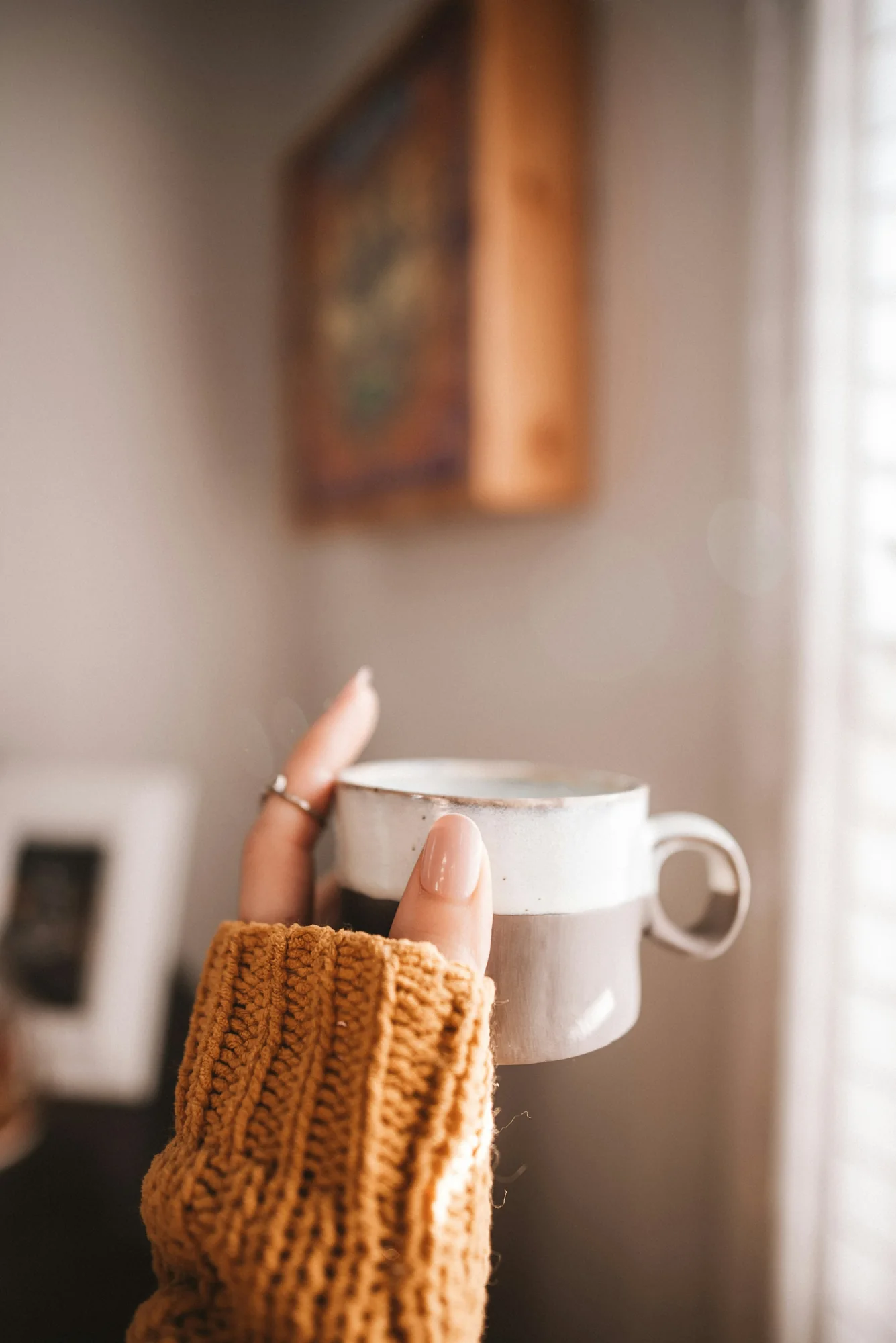 Hand with a glossy nude almond manicure and a mustard knit sleeve holding a speckled stoneware mug