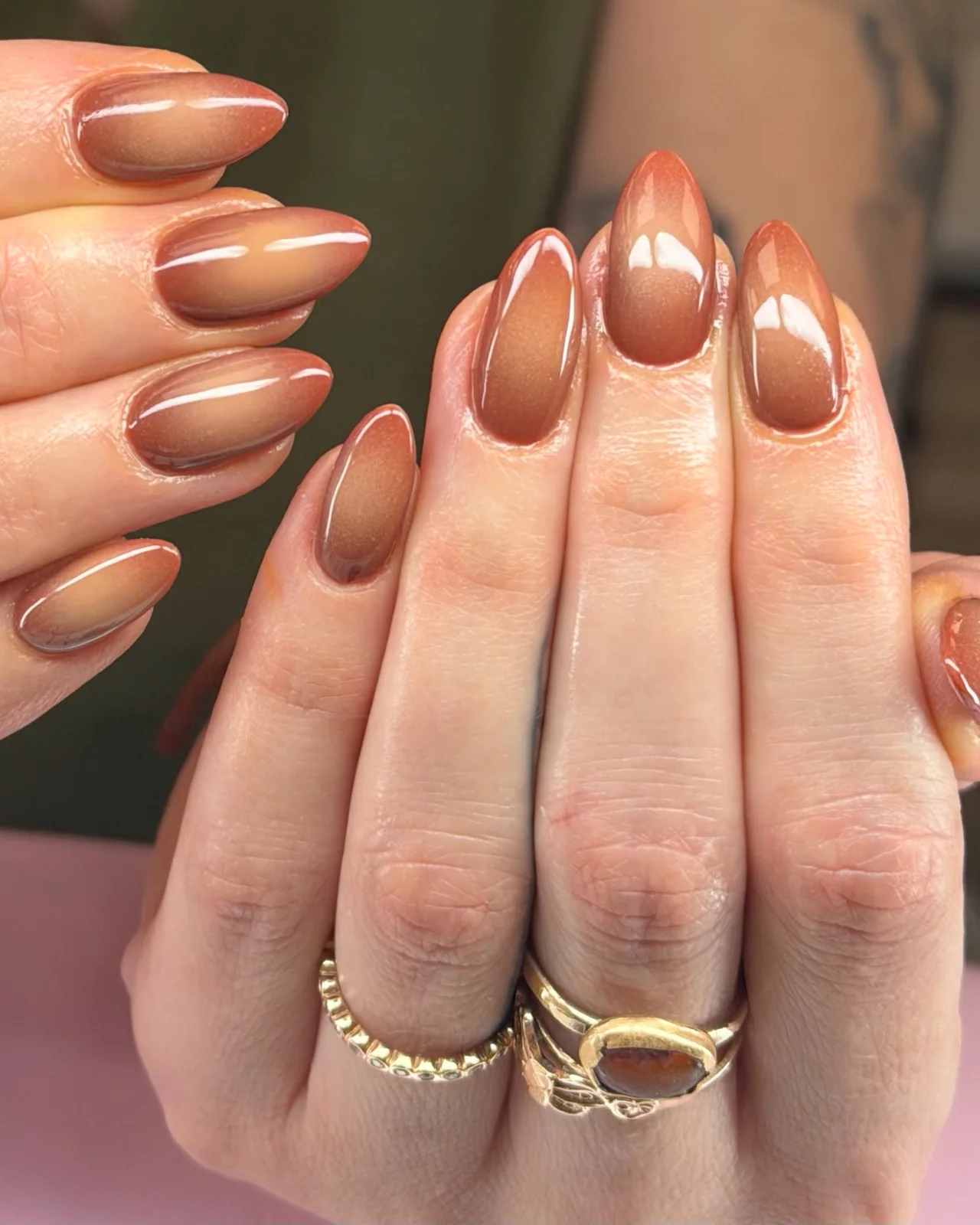 Almond nails in a glossy brown ombré with a fine shimmer, a hand with gold rings
