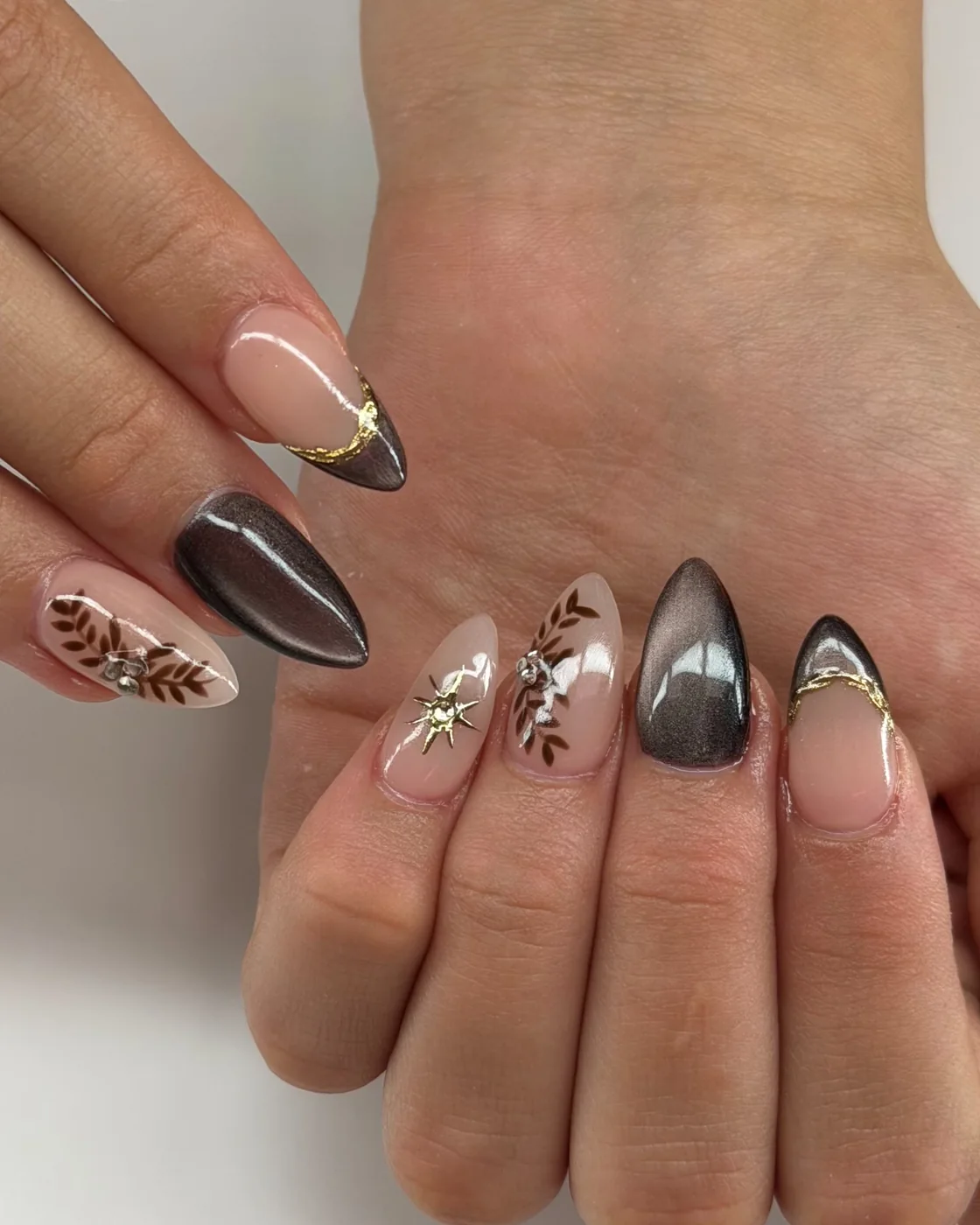 Almond nails in espresso-brown cat-eye, nude with cat-eye French tips edged in gold foil, brown hand-painted leaves with rhinestones, and a gold star stud