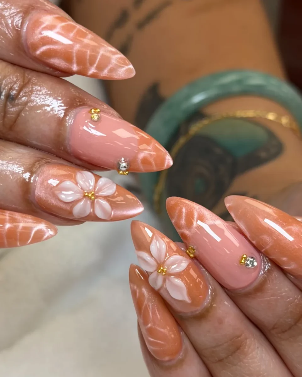 Long stiletto nails in peach shimmer with white ripple lines, white 3D flowers with gold centres, and pink nails with gold beads and a rhinestone