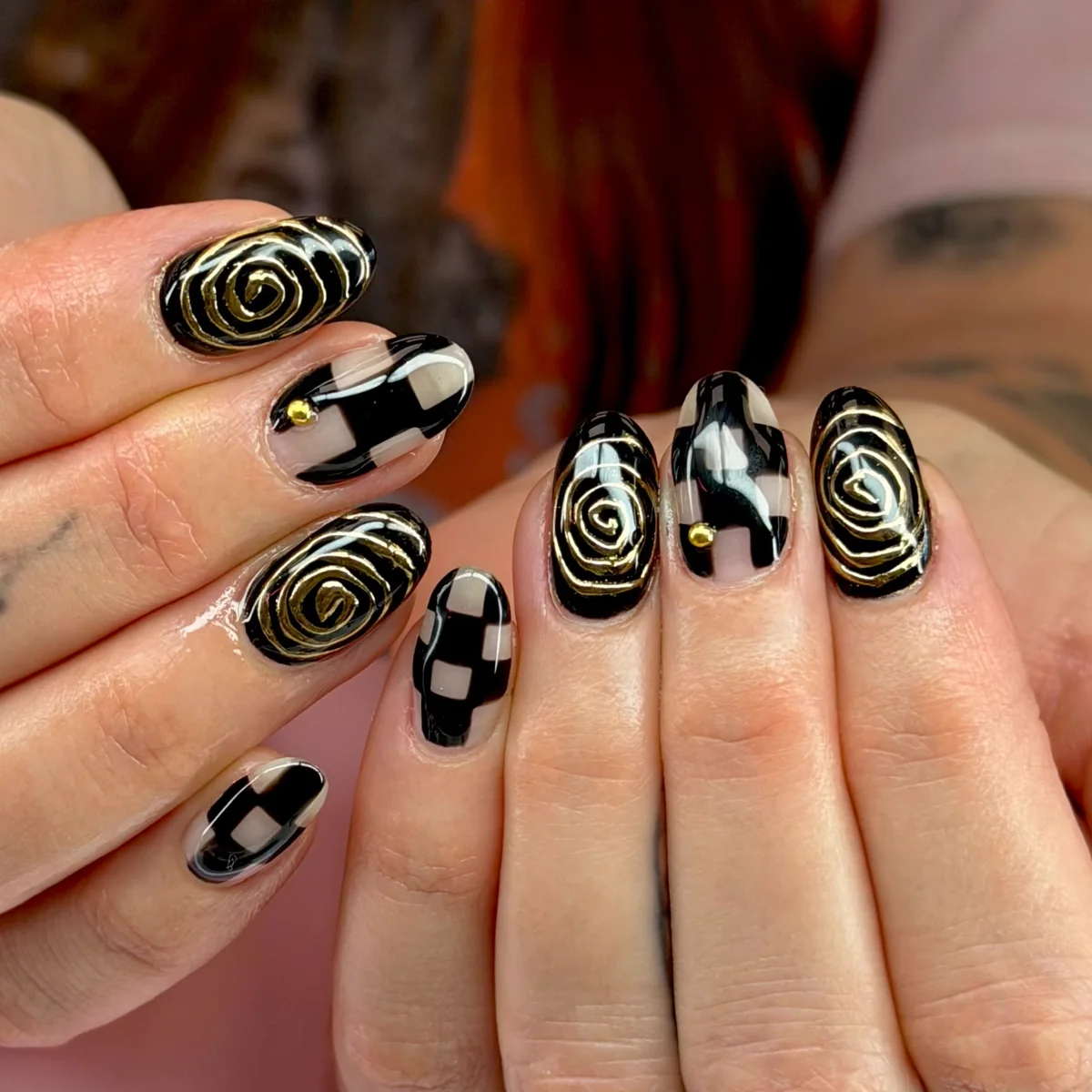 Short almond nails in glossy black with raised gold spirals, and black-and-nude checkerboard with a small gold stud