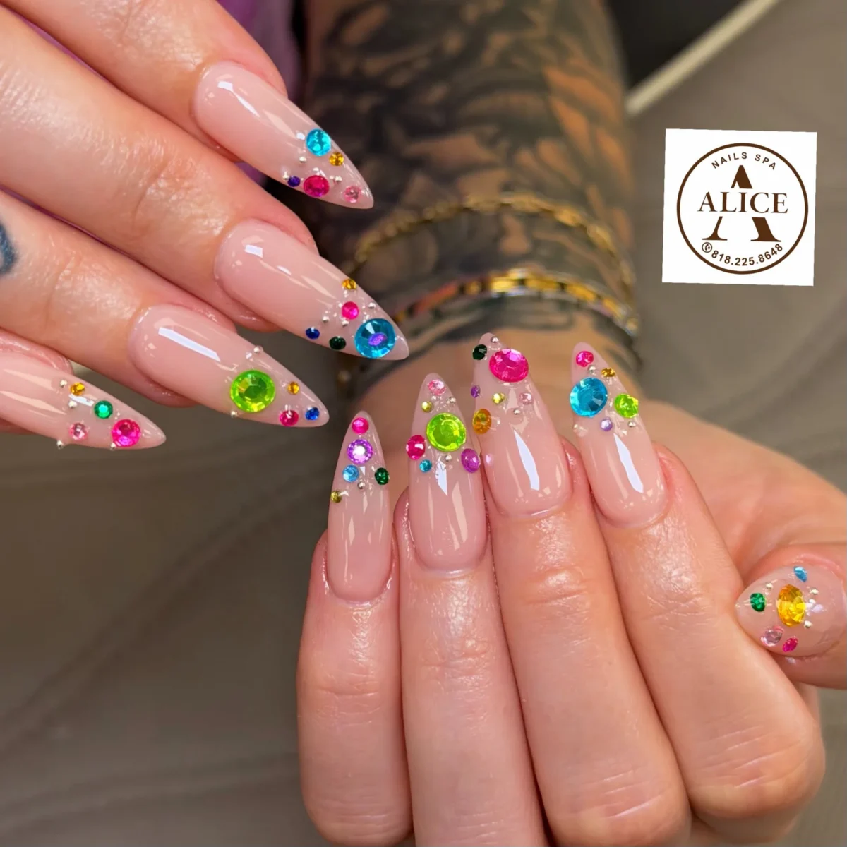 Long nude stiletto nails with clusters of bright rhinestones in pink, green, blue, purple and orange near the tips