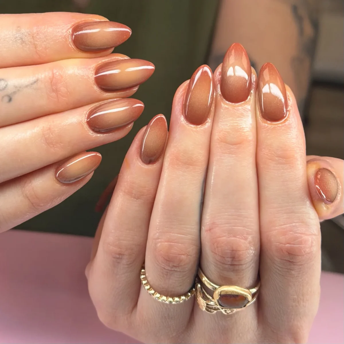 Almond nails in a glossy brown ombré with a fine shimmer, a hand with gold rings