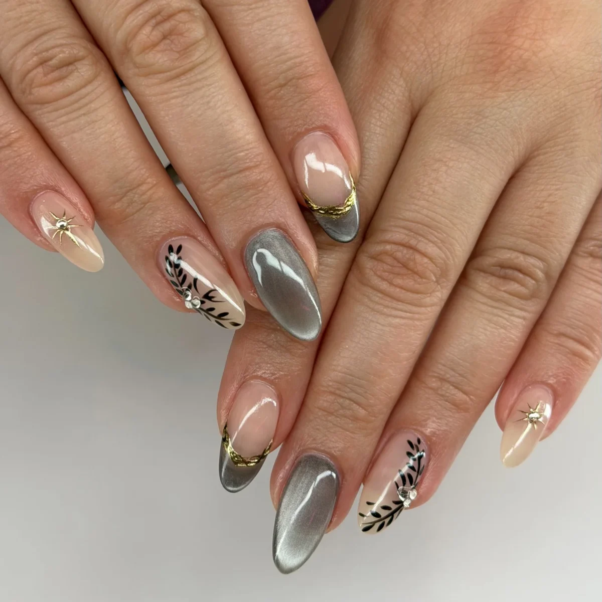 Almond nails in grey cat-eye, nude with cat-eye French tips edged in gold foil, black hand-painted leaves with rhinestones, and gold star studs