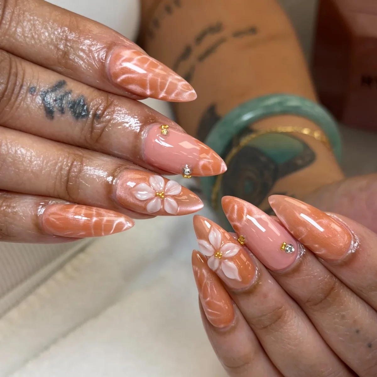 Long stiletto nails in peach shimmer with white ripple lines, white 3D flowers with gold centres, and pink nails with gold beads and a rhinestone