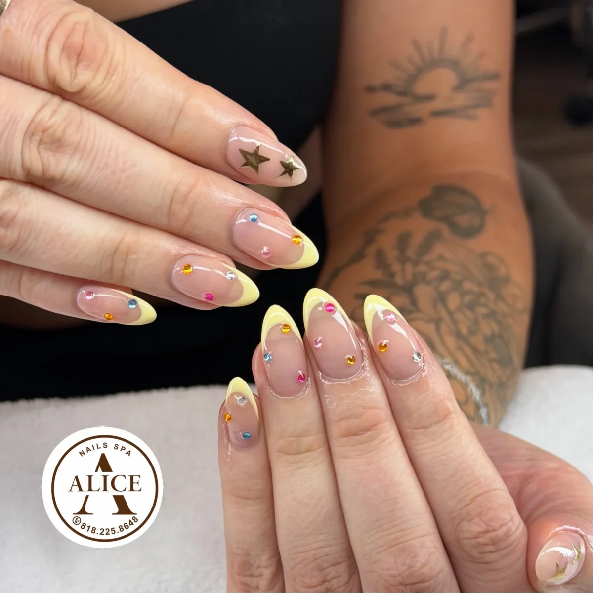 Almond nails with pastel yellow French tips, small rhinestones in pink, orange, blue and clear, and two gold stars on a sheer nude base