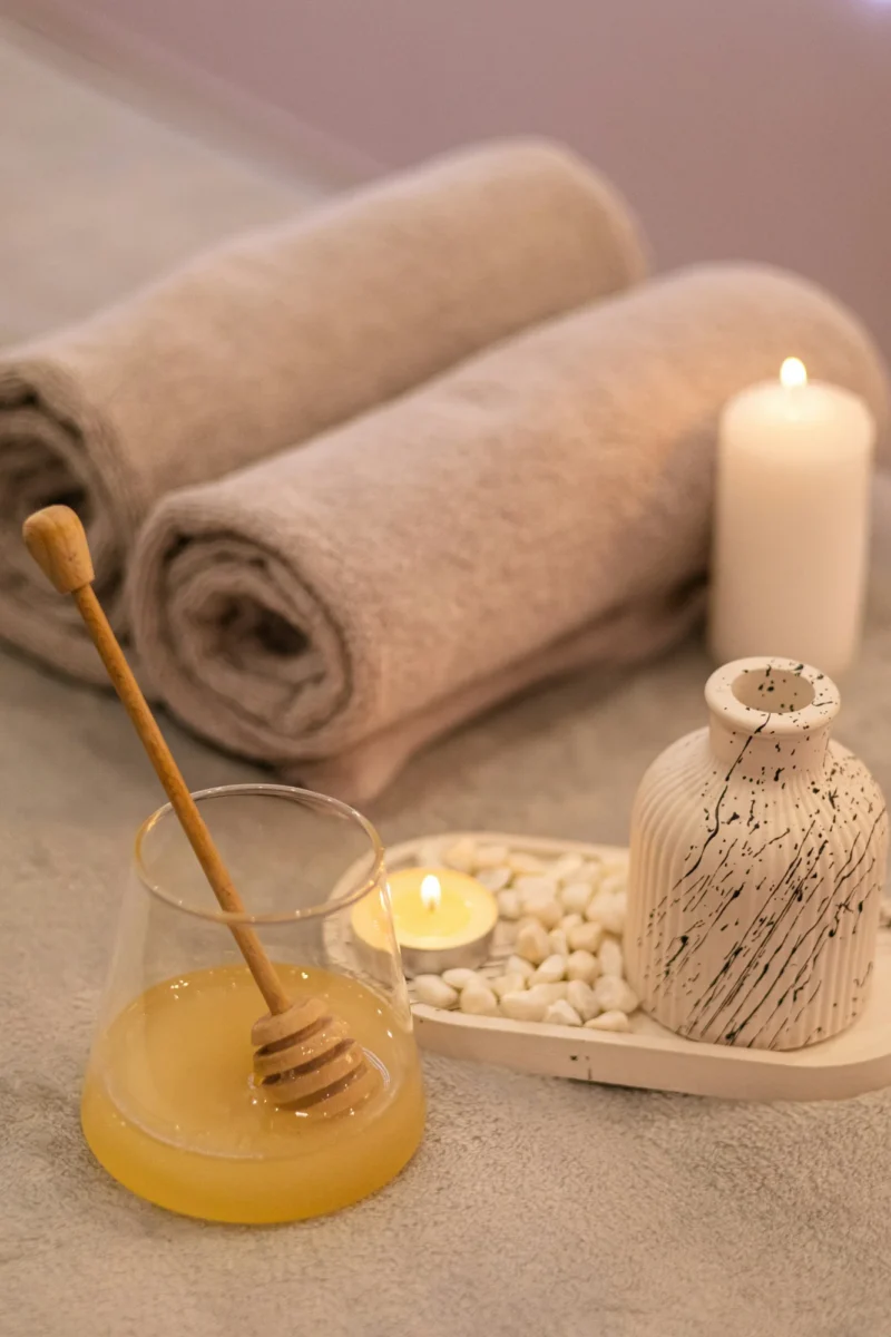 Glass of golden honey with a wooden dipper beside a lit tea light, white pebbles and rolled taupe towels